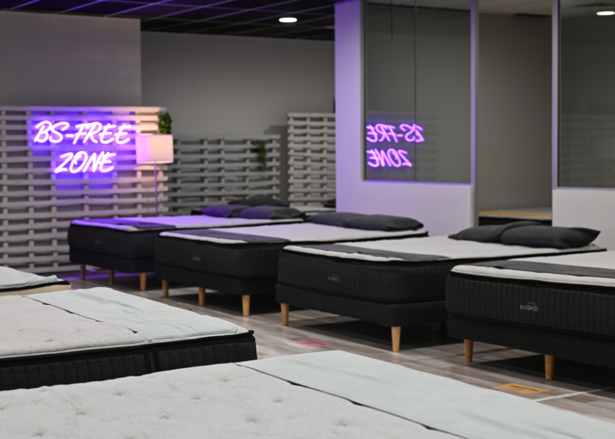 Interior of the Ausbeds Marrickville showroom. Multiple mattresses on wooden leg bases, purple BS-FREE ZONE neon sign on slatted wall, reflected in glass. Moody purple lighting.