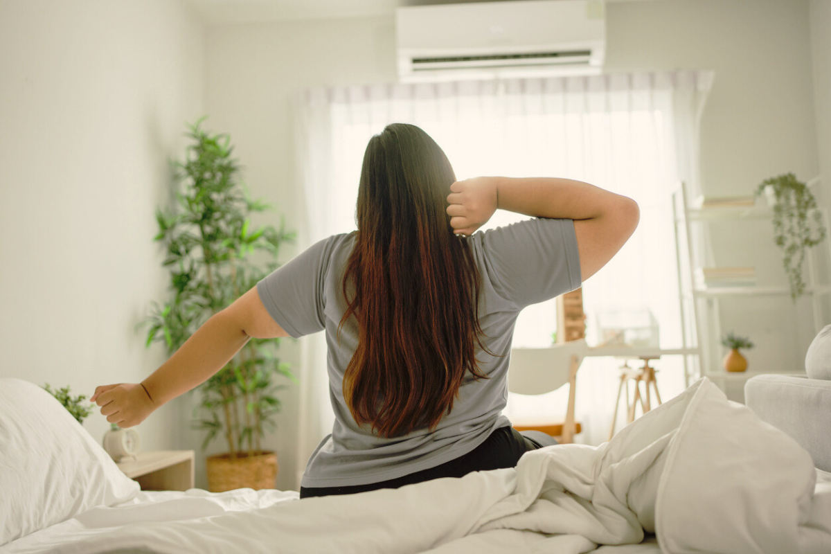A plus-sized person stretches while sitting on a bed in a softly lit bedroom. 